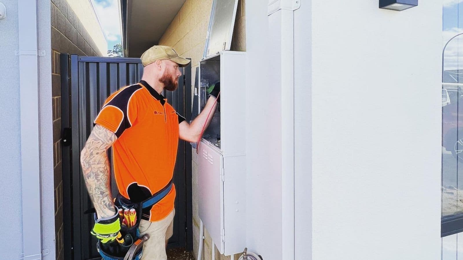 A PSW technician in an orange work polo and tool belt working inside an open outdoor meter box beside a house