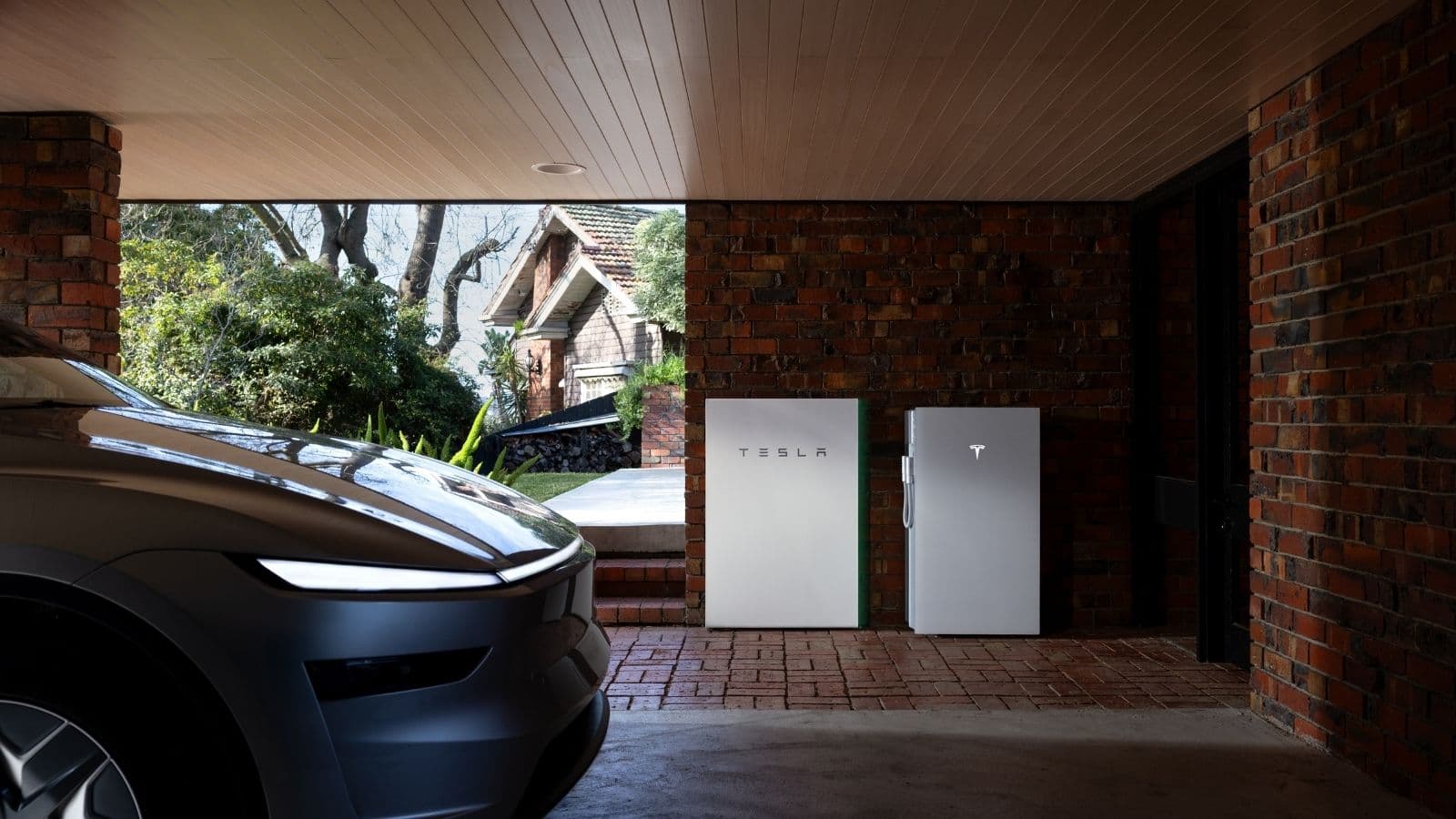 Two Tesla Powerwall units mounted side by side on a brick wall in a carport, with a vehicle parked alongside